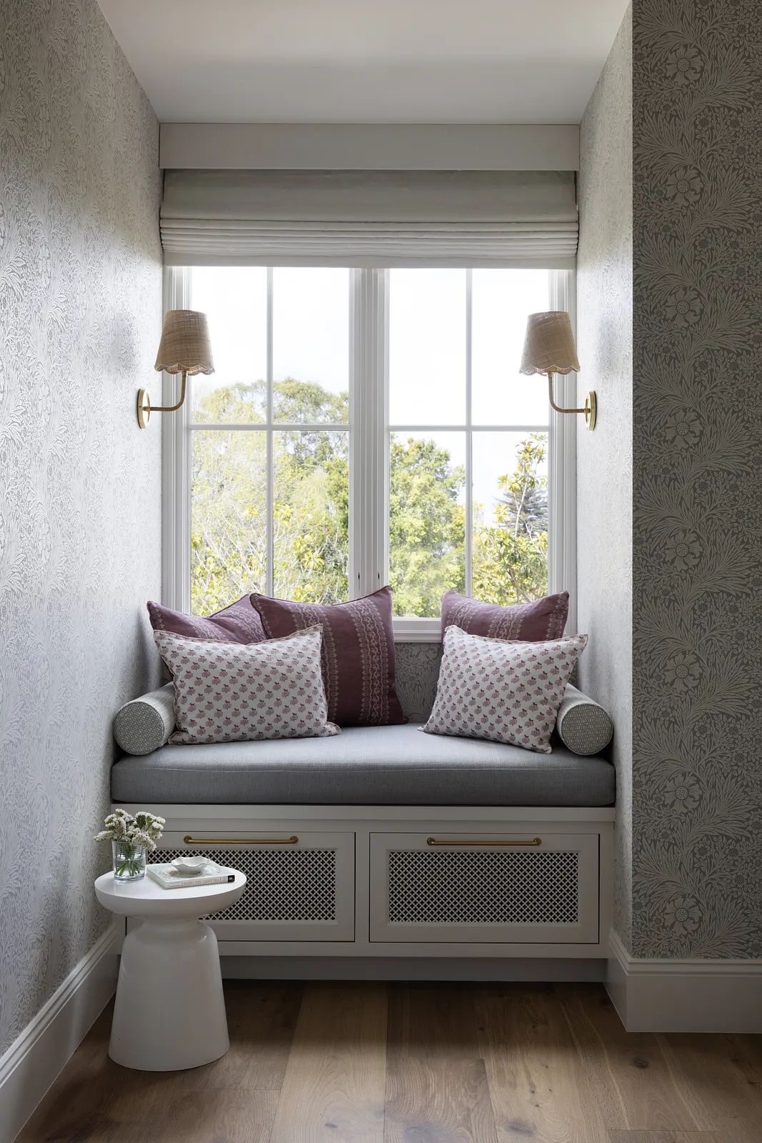 Seating nook with built-in bench, decorative pillows, side table, large window, and patterned wallpaper.