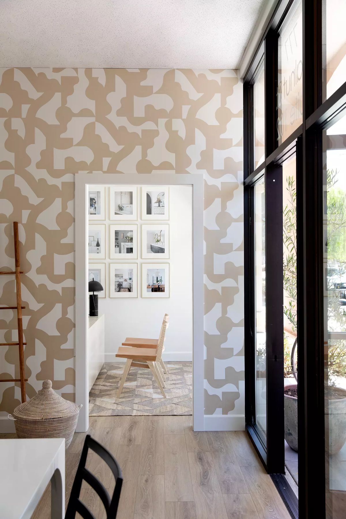 Entryway features patterned wallpaper, wooden ladder, gallery wall, chair, and large glass doors with plants.