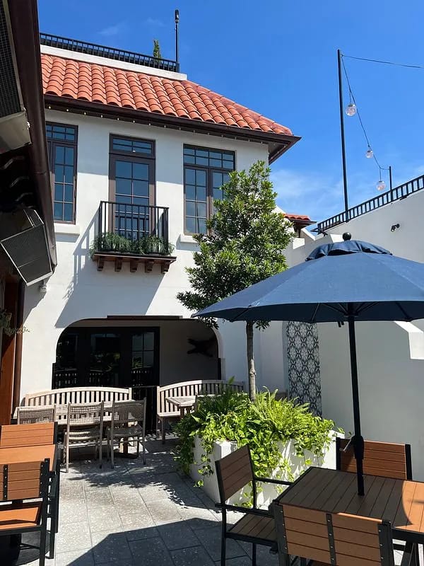 Patio with wooden tables, chairs, umbrella, potted plants, and a background building with red tile roof.