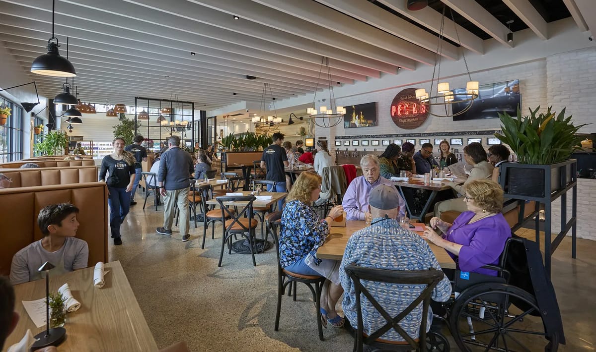 Dining room with wooden tables, chairs, patrons, plants, large windows, and exposed beams in the ceiling.