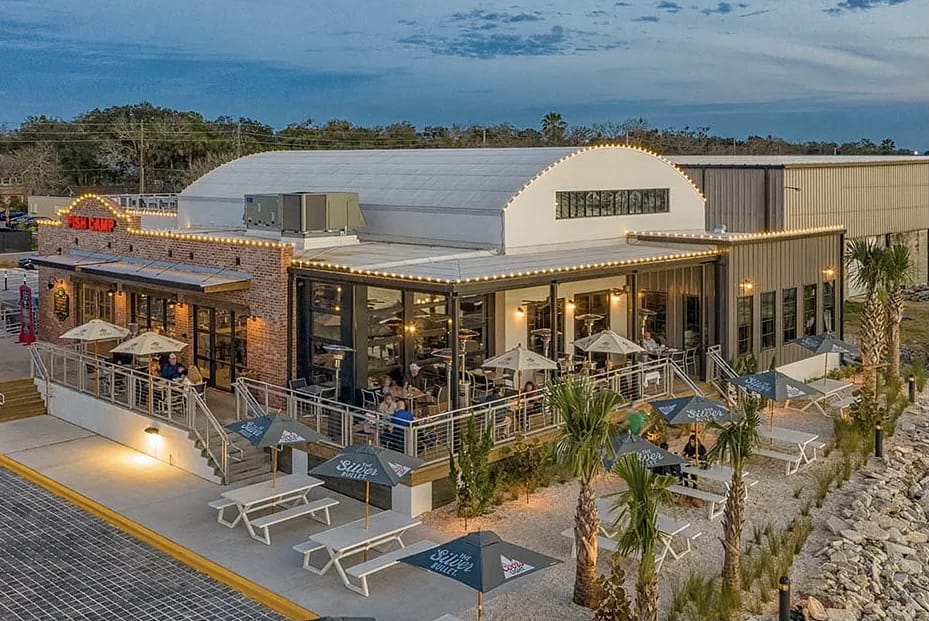Restaurant exterior with modern facade, large windows, outdoor seating, and palm trees.
