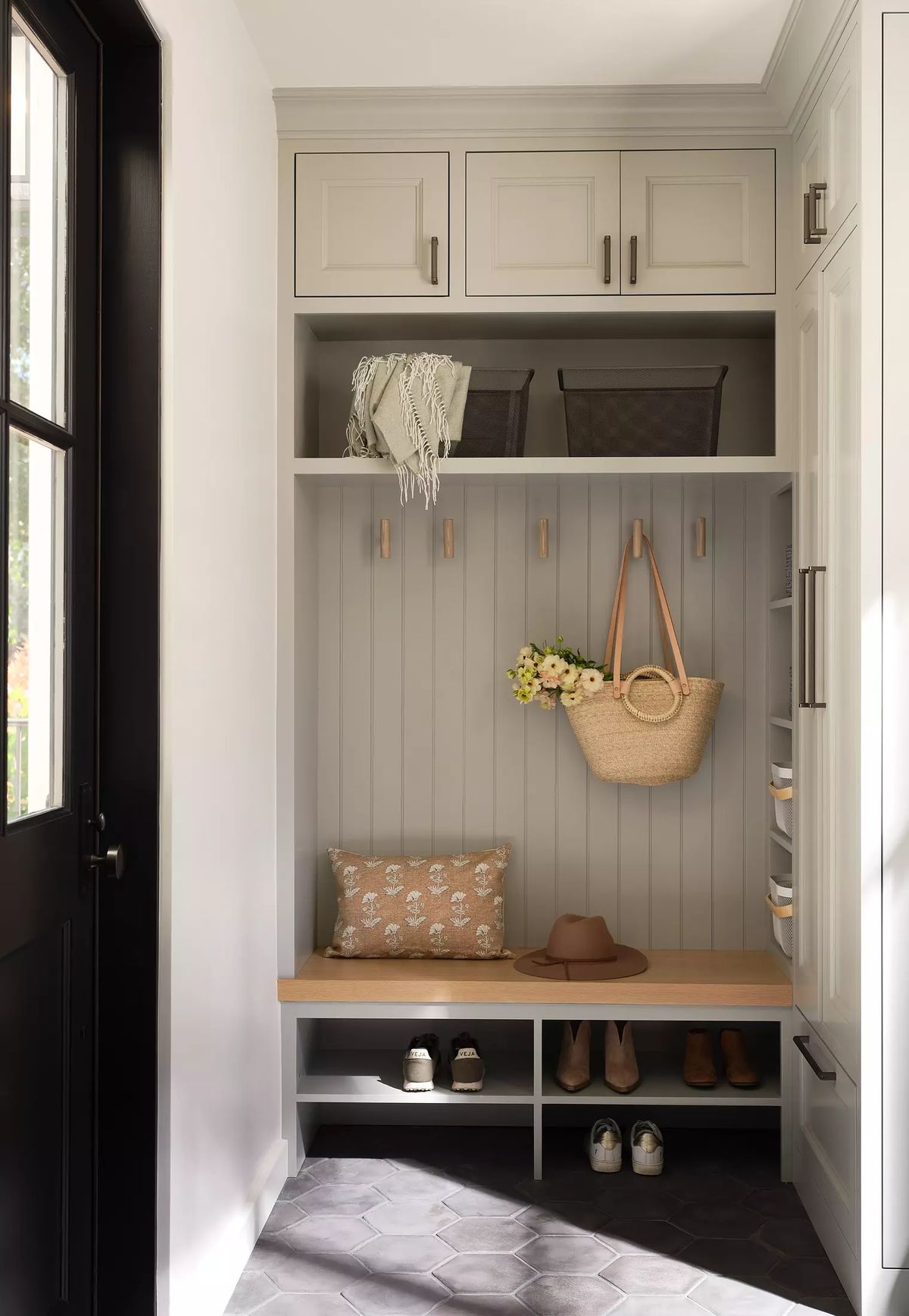 Mudroom with gray bench, upper cabinets, hooks, decorative pillow, shoes, and dark hexagonal tile flooring.