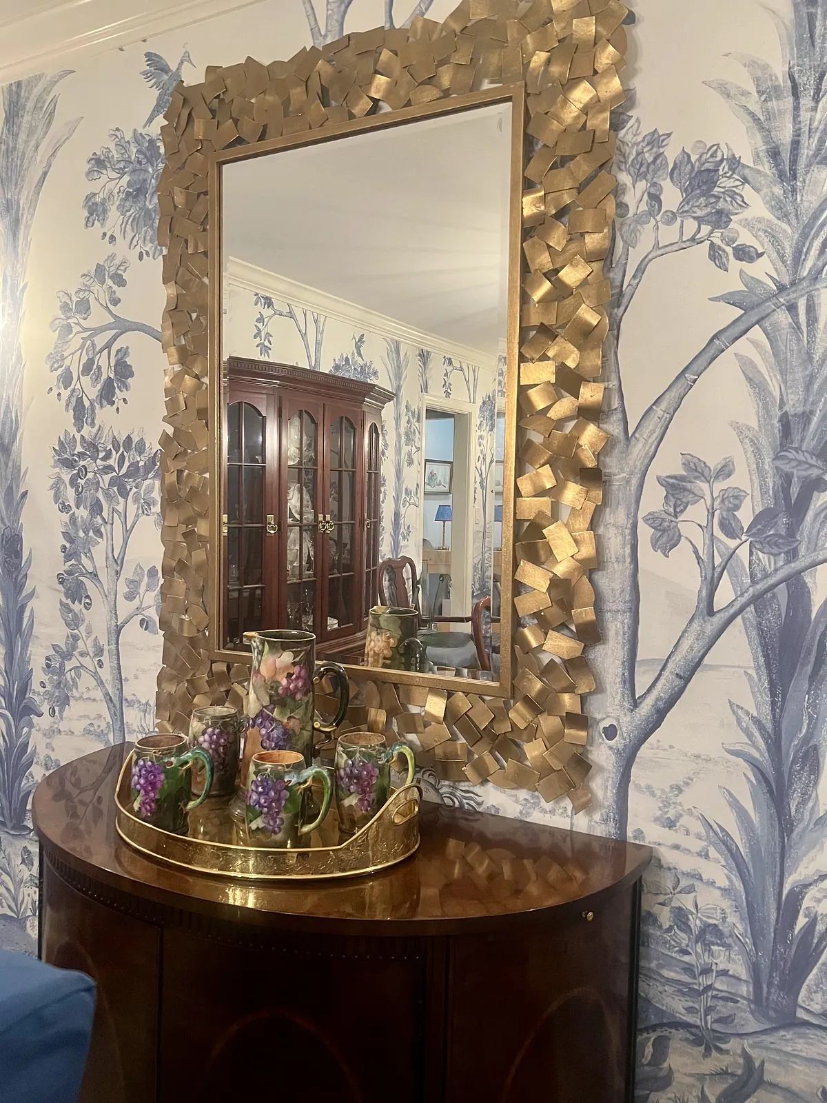 Foyer with gold-framed mirror, wooden console, floral mugs, and patterned wallpaper.