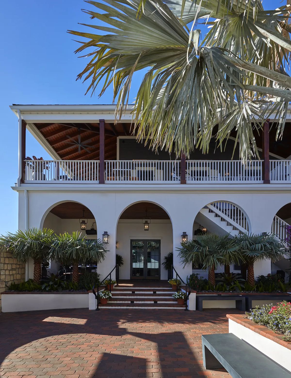 Front exterior with arched doorways, balcony, ceiling fans, brick paving, and tropical landscaping.