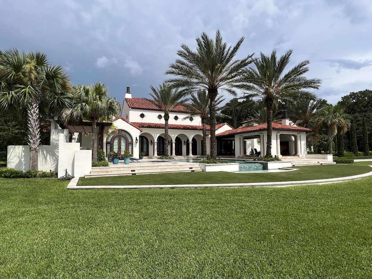 Exterior front view of a house with red-tiled roof, white walls, arched windows, and surrounding palm trees.