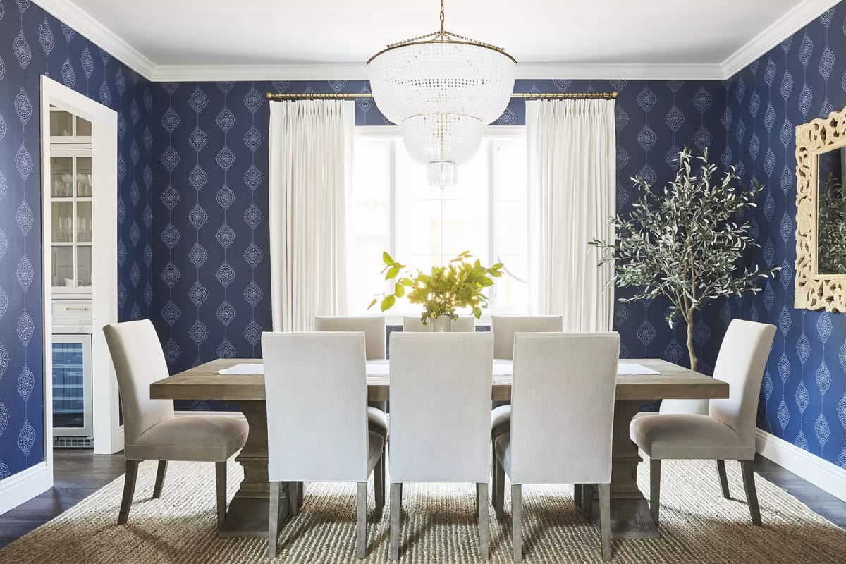 Dining room with wooden table, eight upholstered chairs, chandelier, blue wallpaper, and natural light from windows.