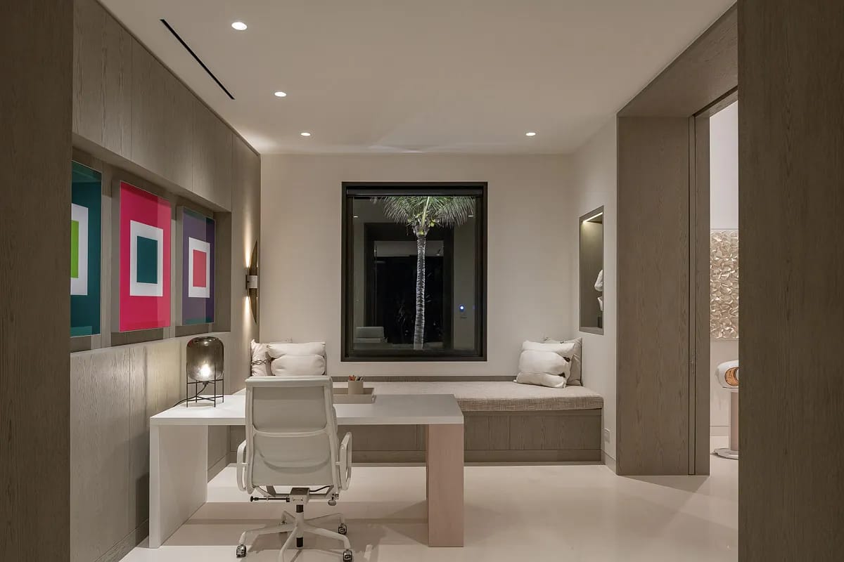 Office with white desk, ergonomic chair, built-in bench, cushions, artwork, and large window.