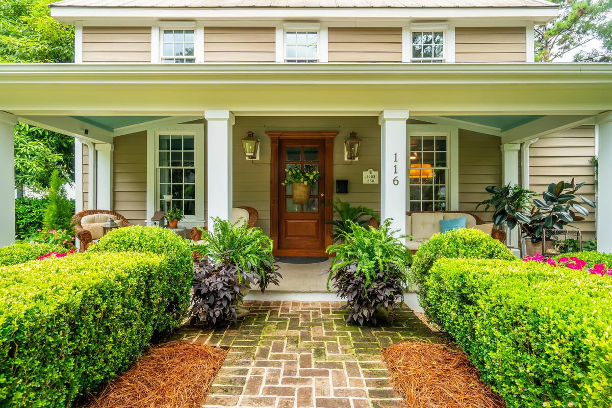 Front exterior with brick walkway, green bushes, and armchairs beside the wooden entrance door.