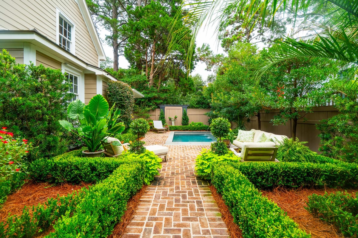 Garden with swimming pool, lounge chairs, and landscaped greenery along a brick pathway.