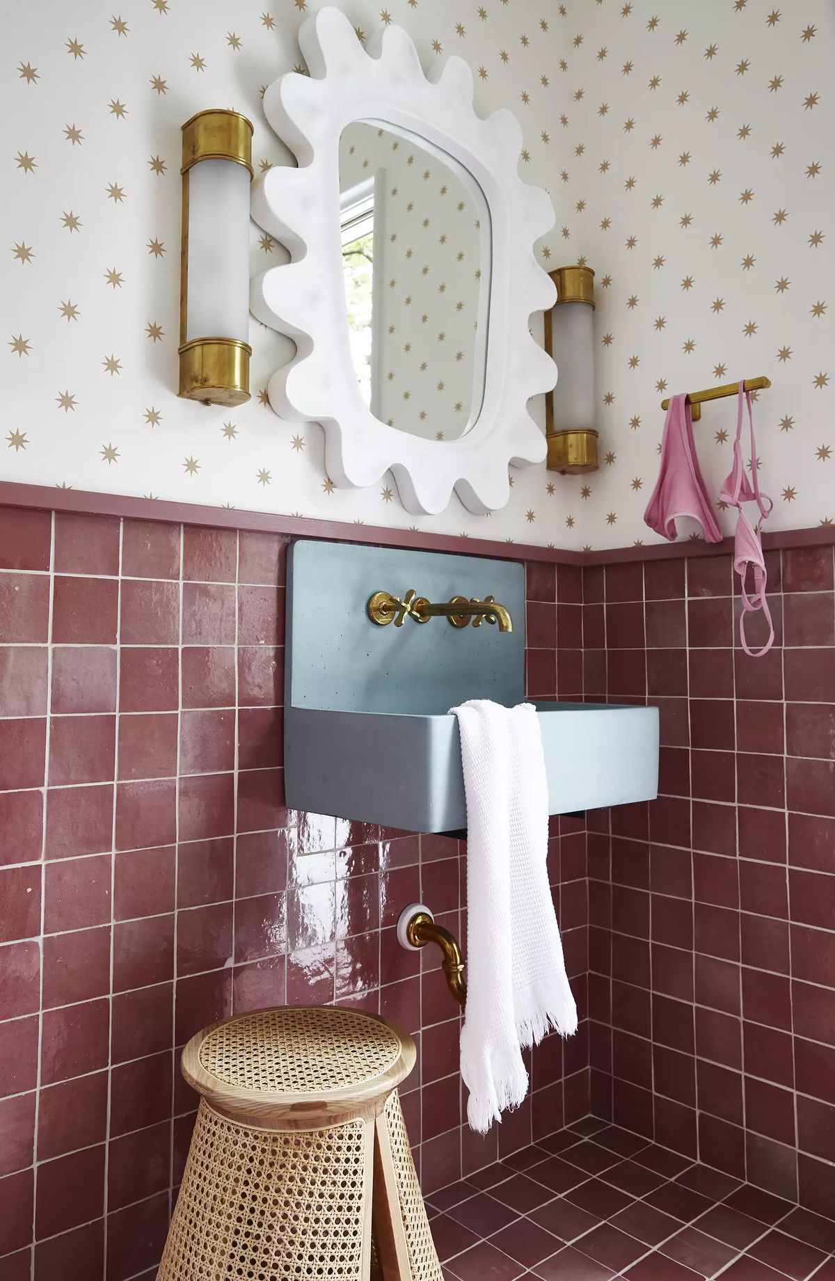Bathroom with wall-mounted sink, gold fixtures, round mirror, pink-patterned walls, and woven stool