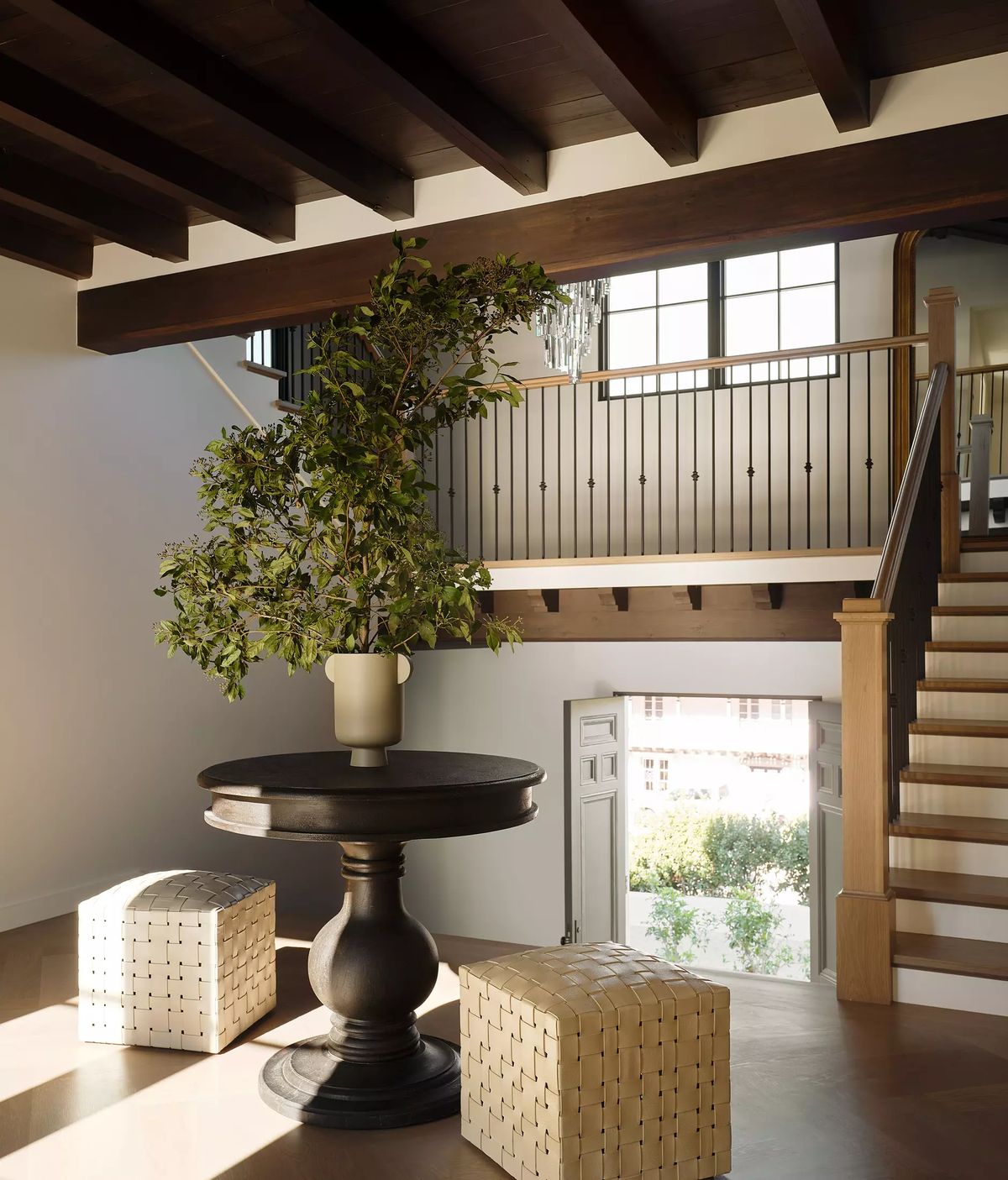 Foyer with round wooden table, potted plant, woven stools, staircase, and large windows for natural light.