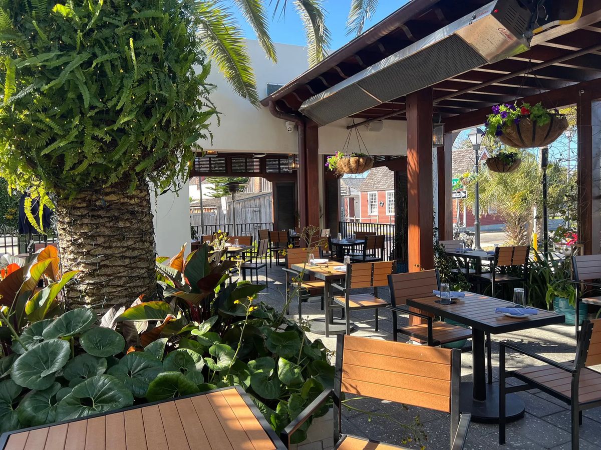 Outdoor patio with wooden tables, chairs, planters, palm tree, and wood beam roof structure.