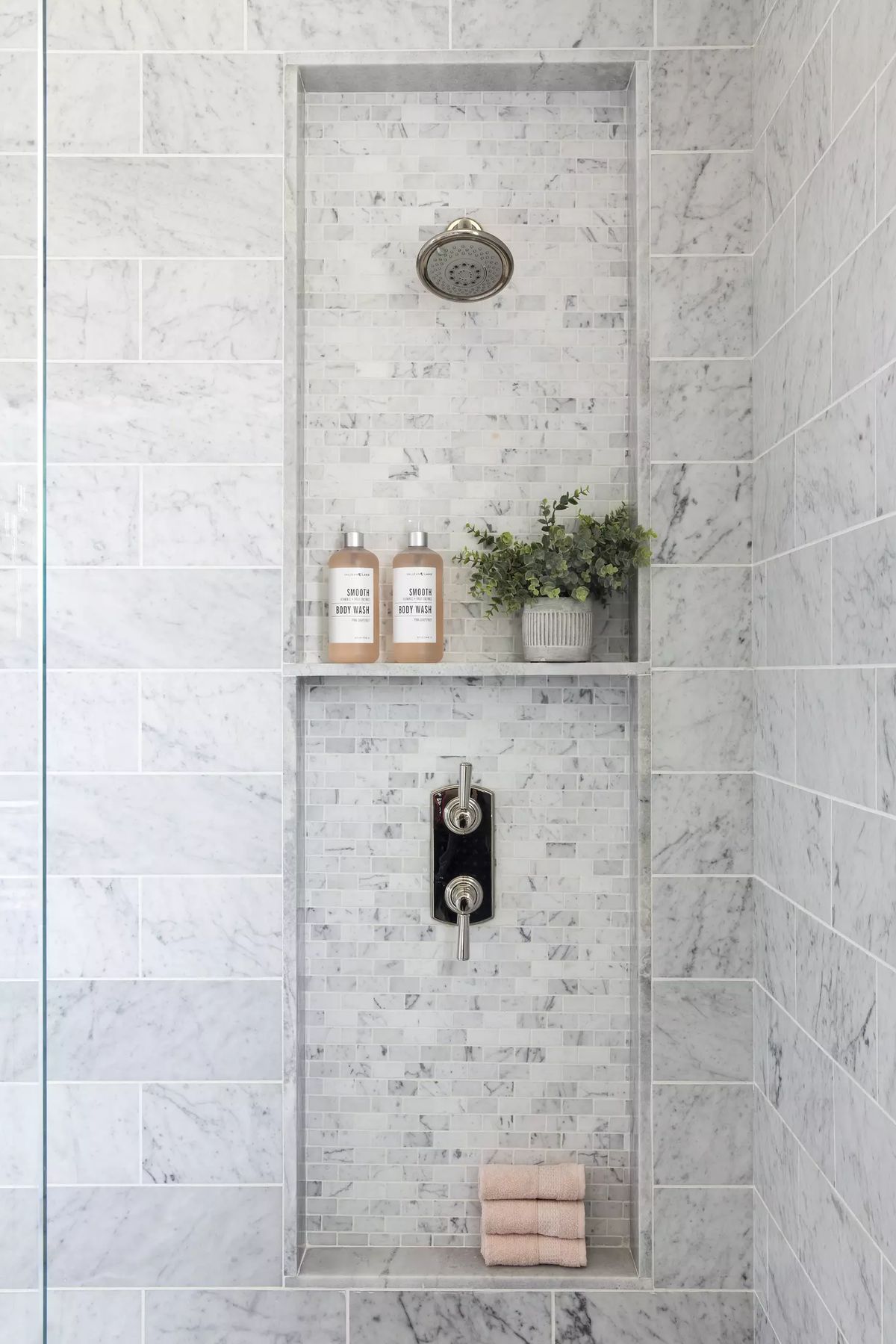 Bathroom shower area with marble tile, built-in niche, shampoo bottles, plant, and pink towel.