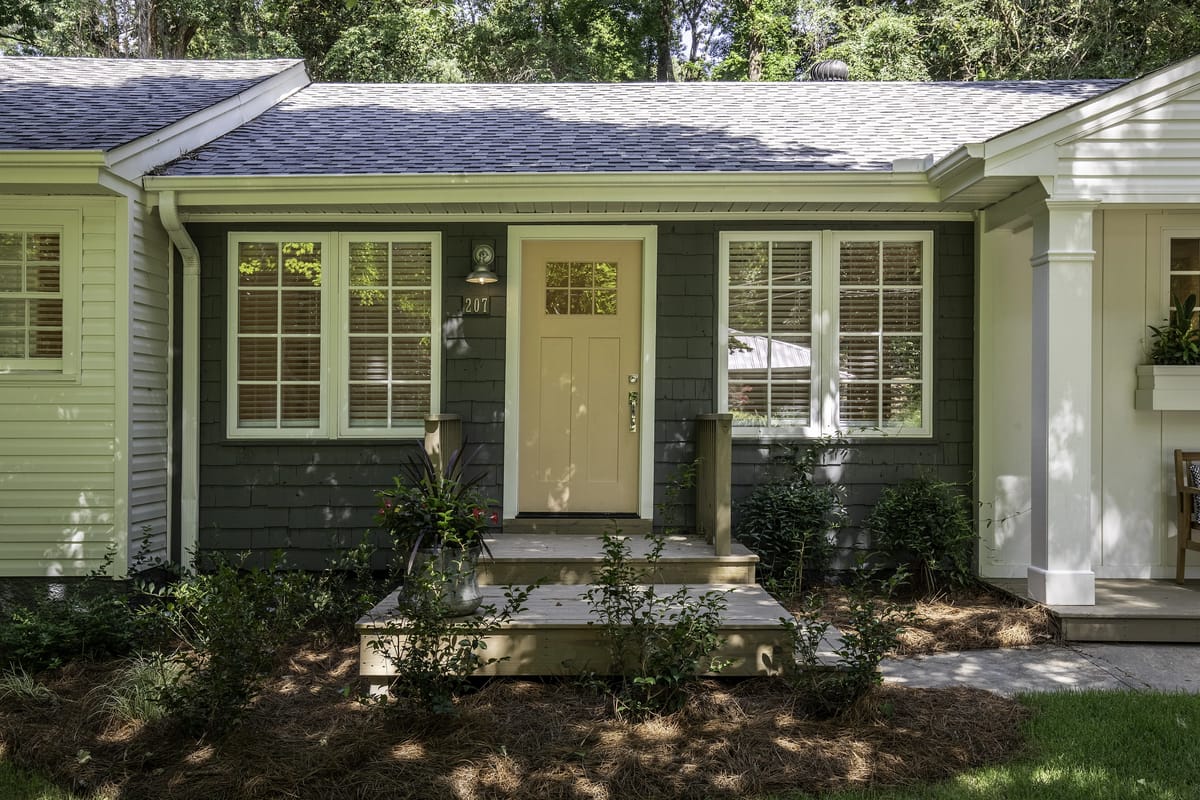 Exterior front with wooden door, six windows, and wooden deck surrounded by shrubs and a garden bed.