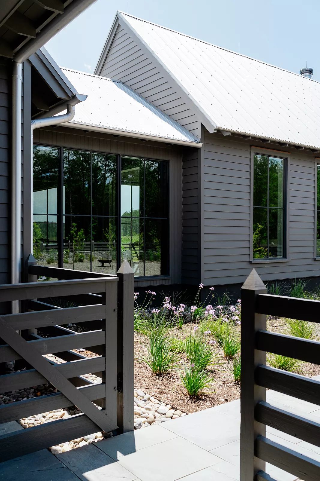 Exterior front of a house with gray facade, large windows, landscaped flower beds, and wooden fence.