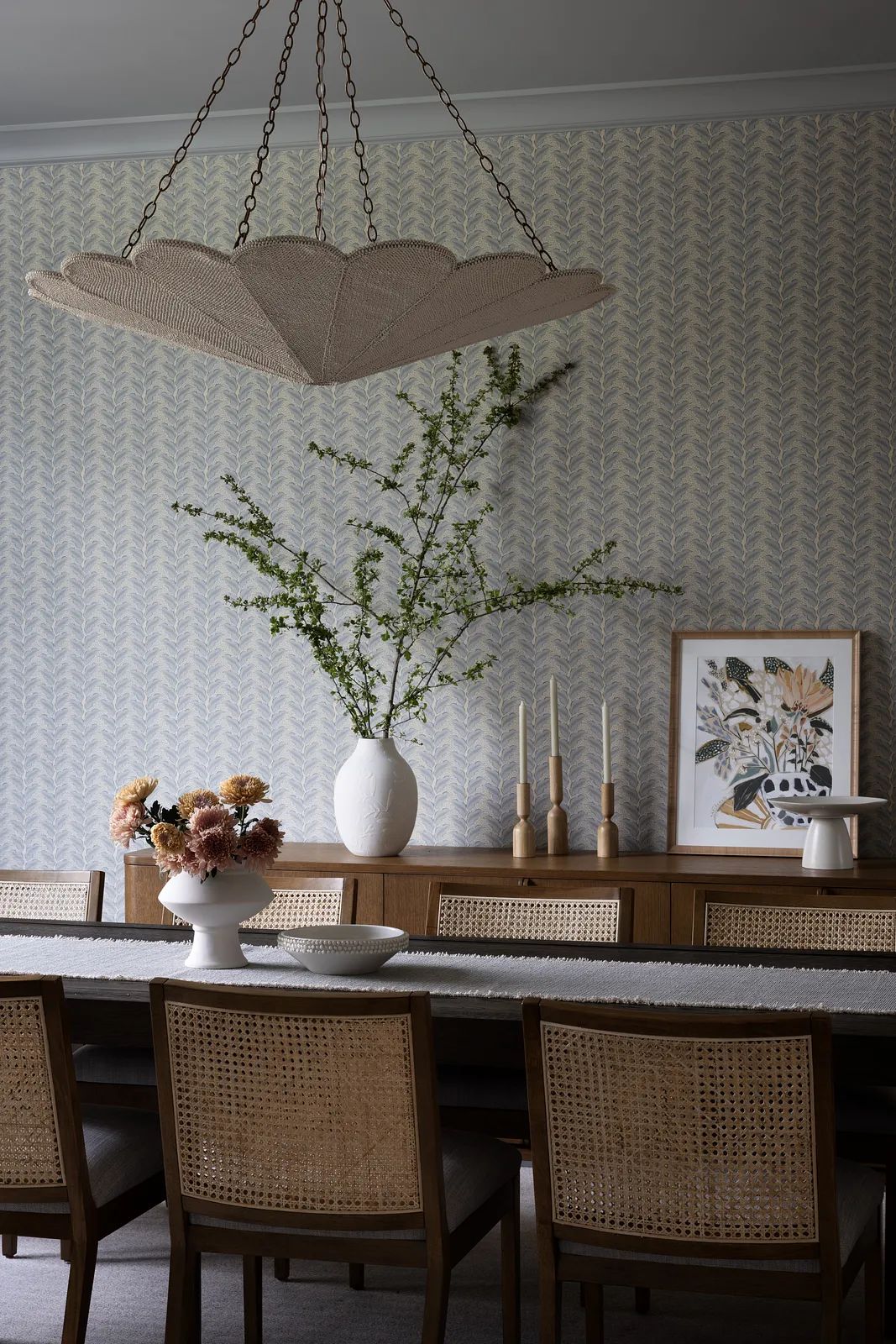 Dining room with long table, caned chairs, flowers, bowl, sideboard, and patterned wallpaper