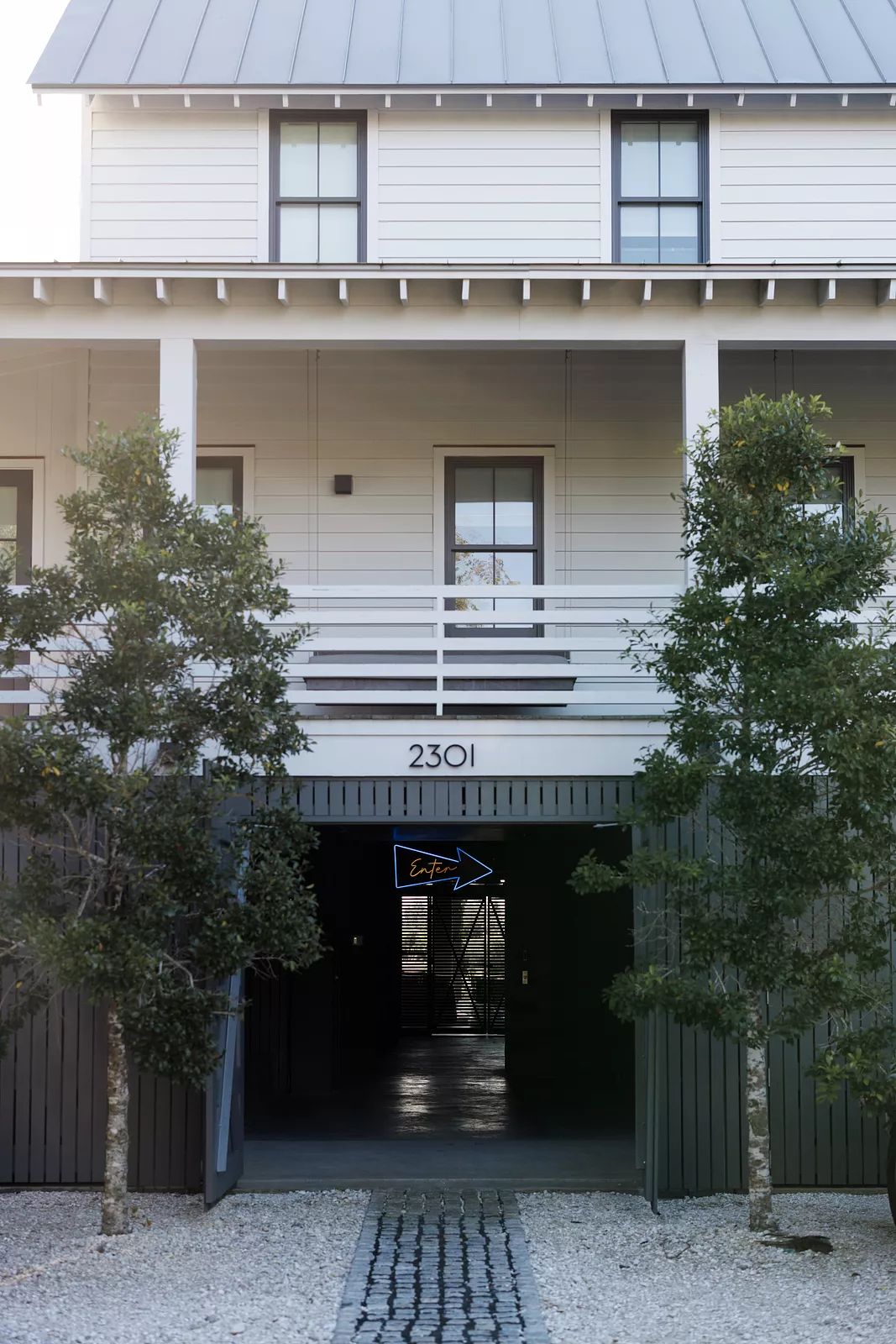 Exterior front view of a building with white siding, metal roof, trees, and gravel pathway.