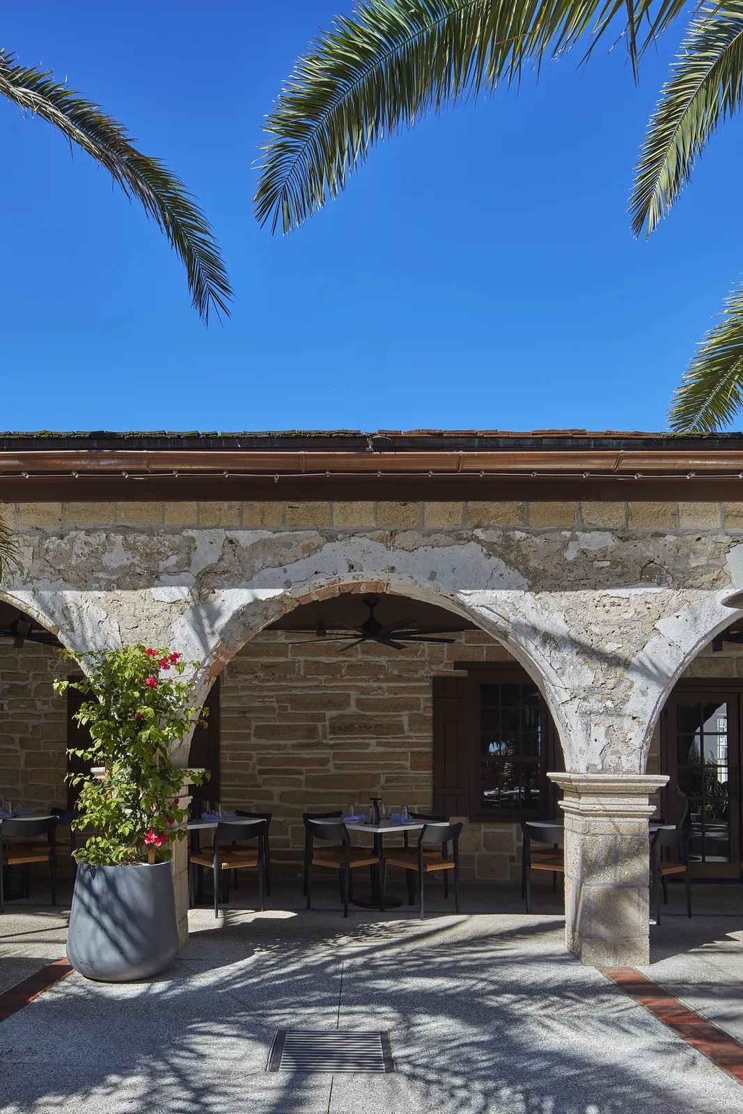 Exterior area with stone arches, palm trees, dining tables, and wooden window shutters.