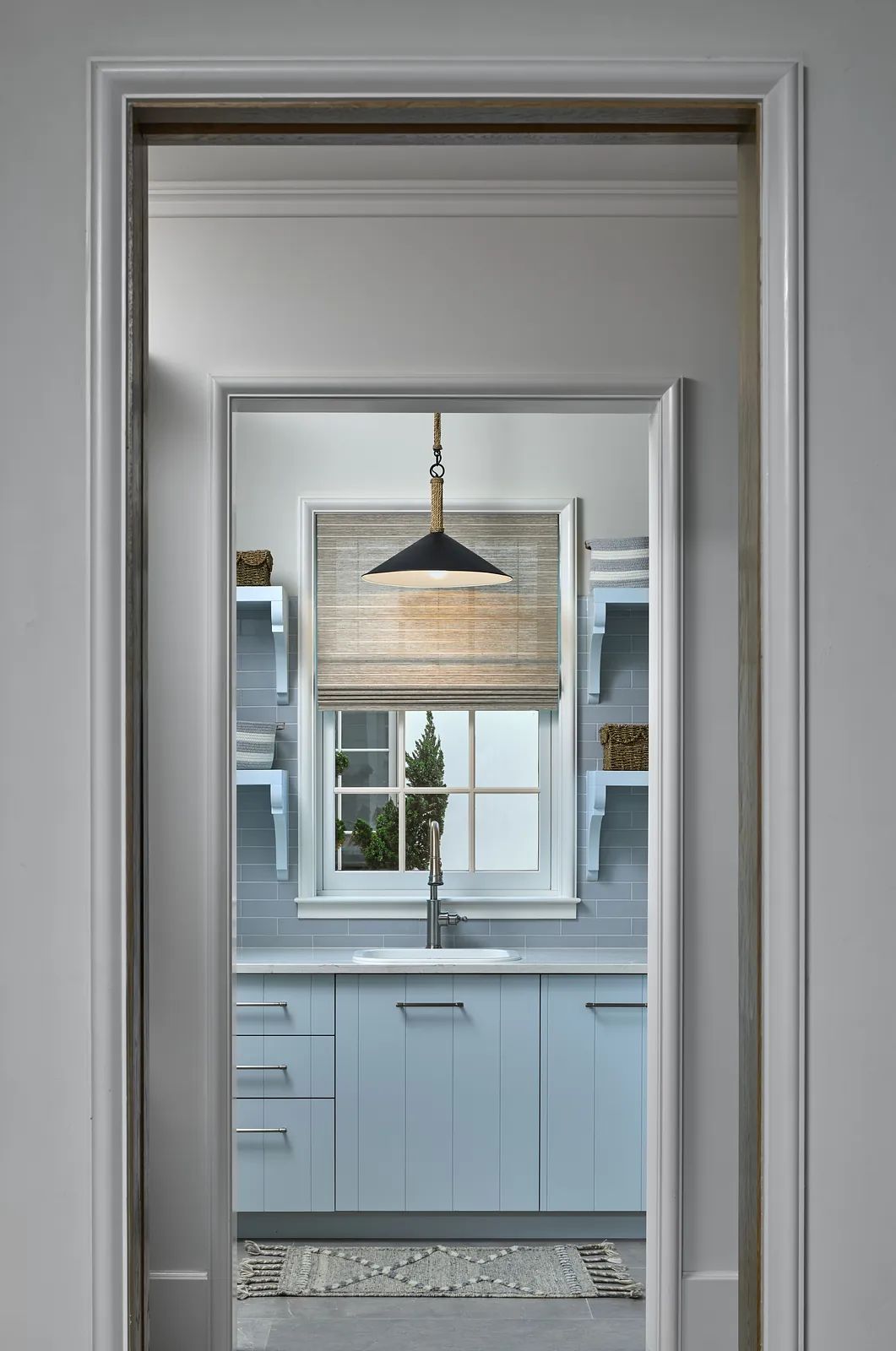 Kitchen with light blue cabinetry, stainless steel sink, window, and pendant light.