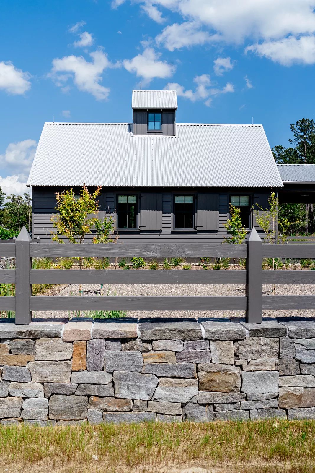 Exterior front view of a gray wooden building with metal roof, windows, stone wall, and landscaping.