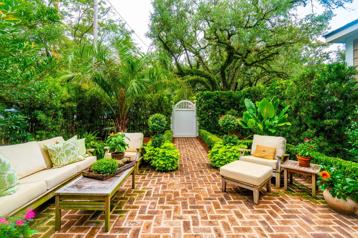 Garden with brick patio, lounge chairs, coffee table, and various potted plants surrounded by lush greenery