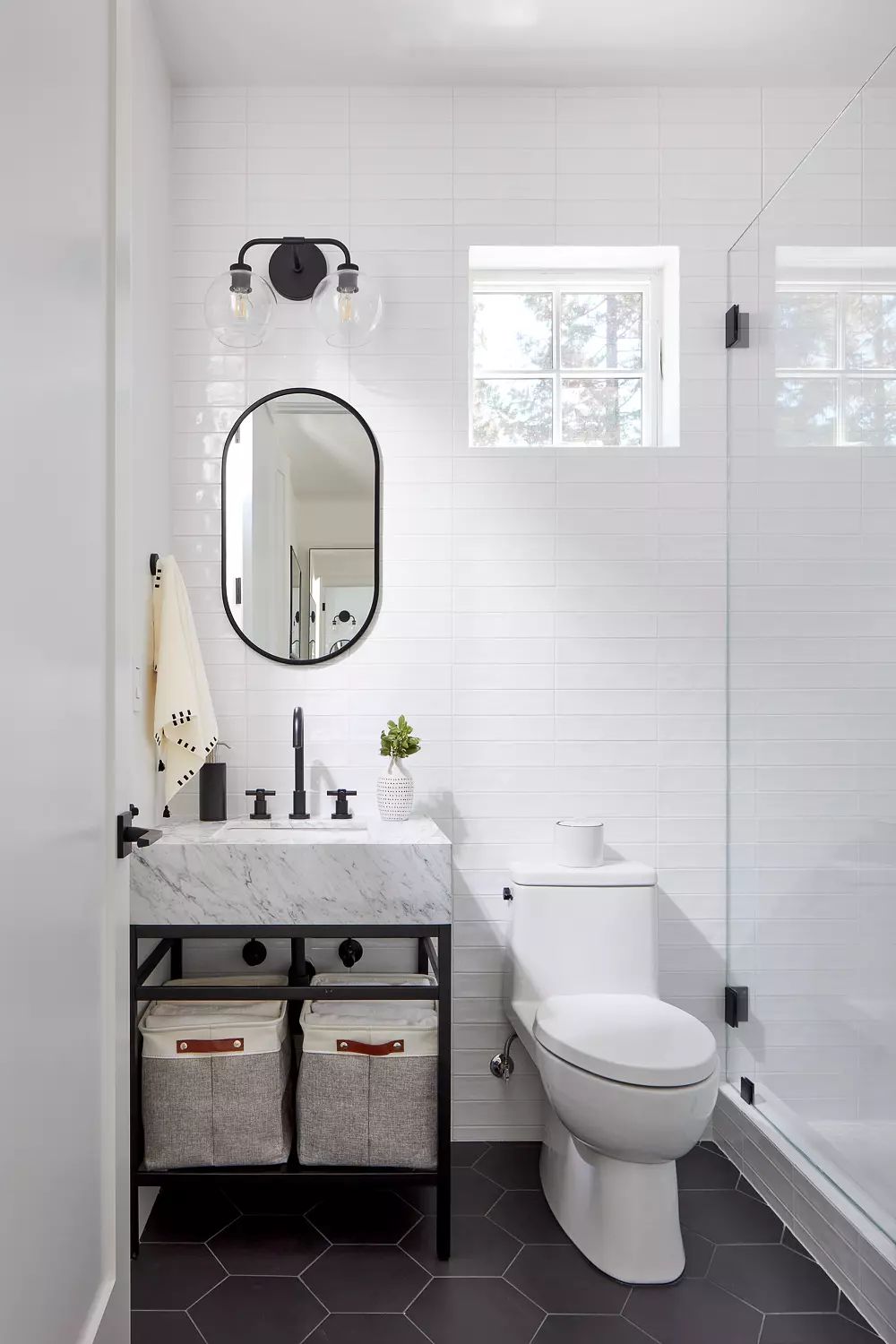 Bathroom with white sink, round mirror, glass shower, toilet, hexagonal black tiles, and a small plant.