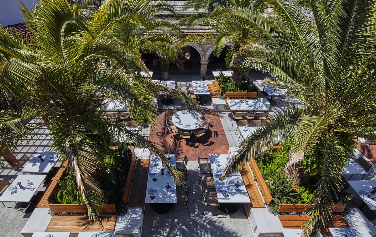 Outdoor patio with wooden tables, white tablecloths, palm trees, and paved flooring.