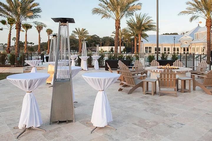 Outdoor patio with round tables, wooden chairs, heating lamps, and central fire pit surrounded by palm trees.