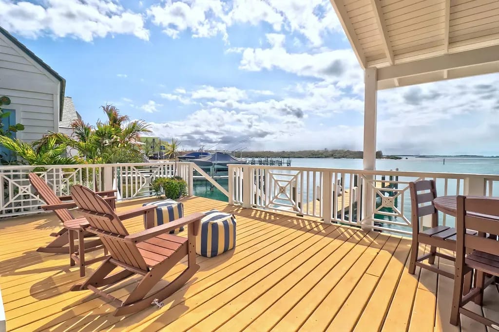 Deck with wooden chairs, striped poufs, round table, and a water view.