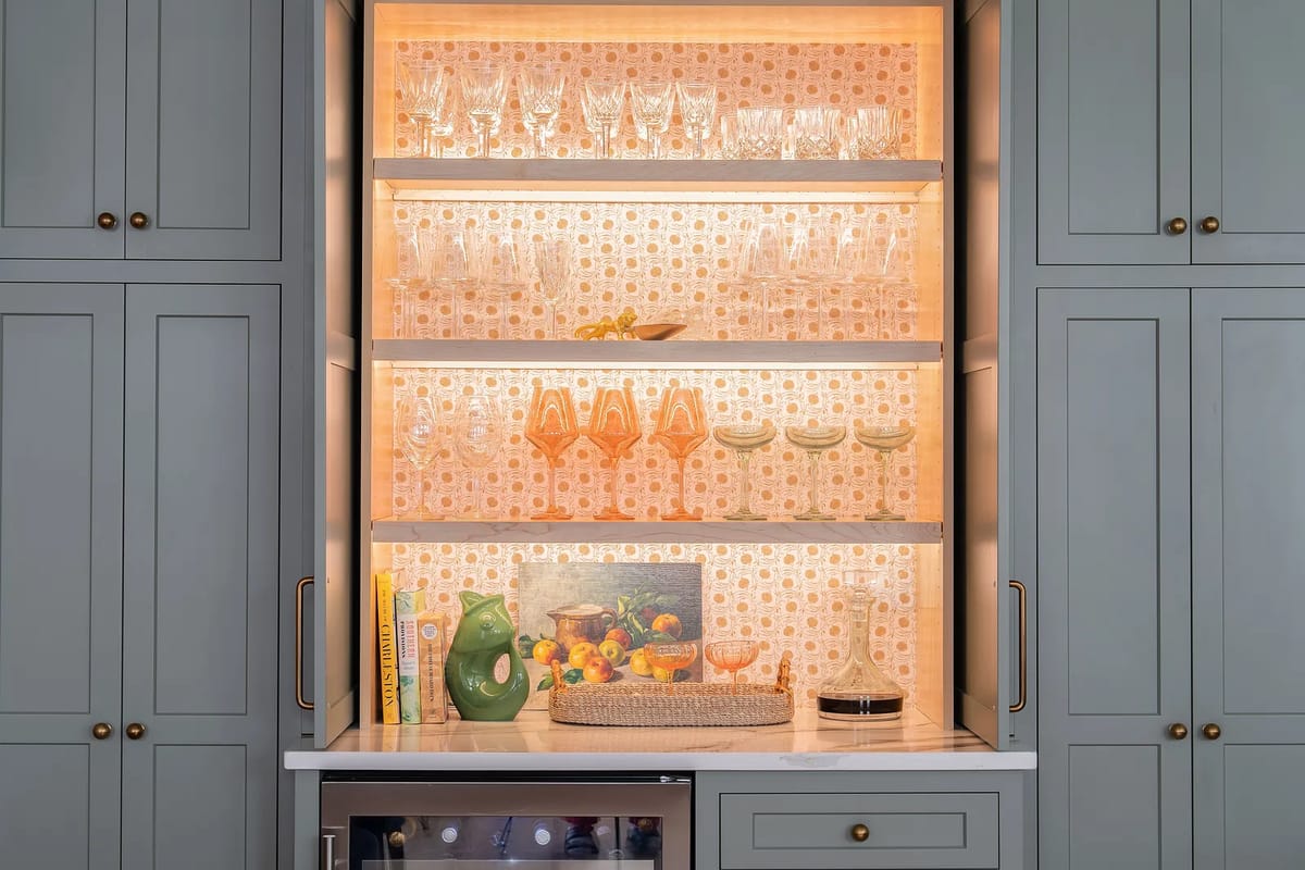 Kitchen with glassware on shelves, gray cabinetry, and countertop items including a green pitcher and decorative basket.