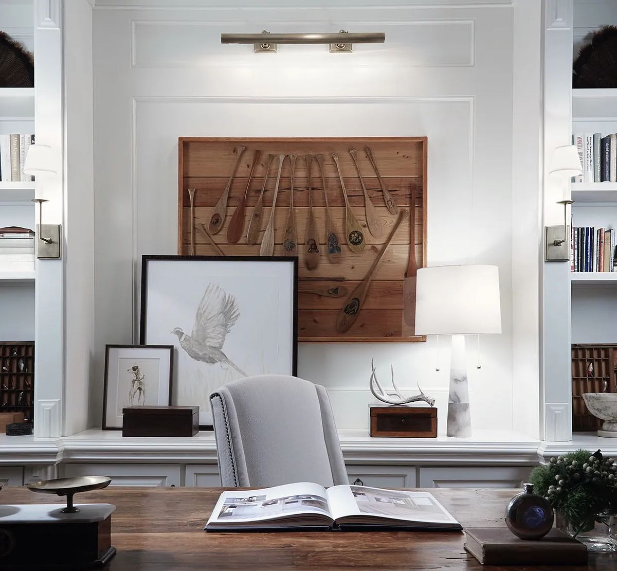 Office with wooden desk, upholstered chair, wall-mounted artwork, bookshelves, and a table lamp.