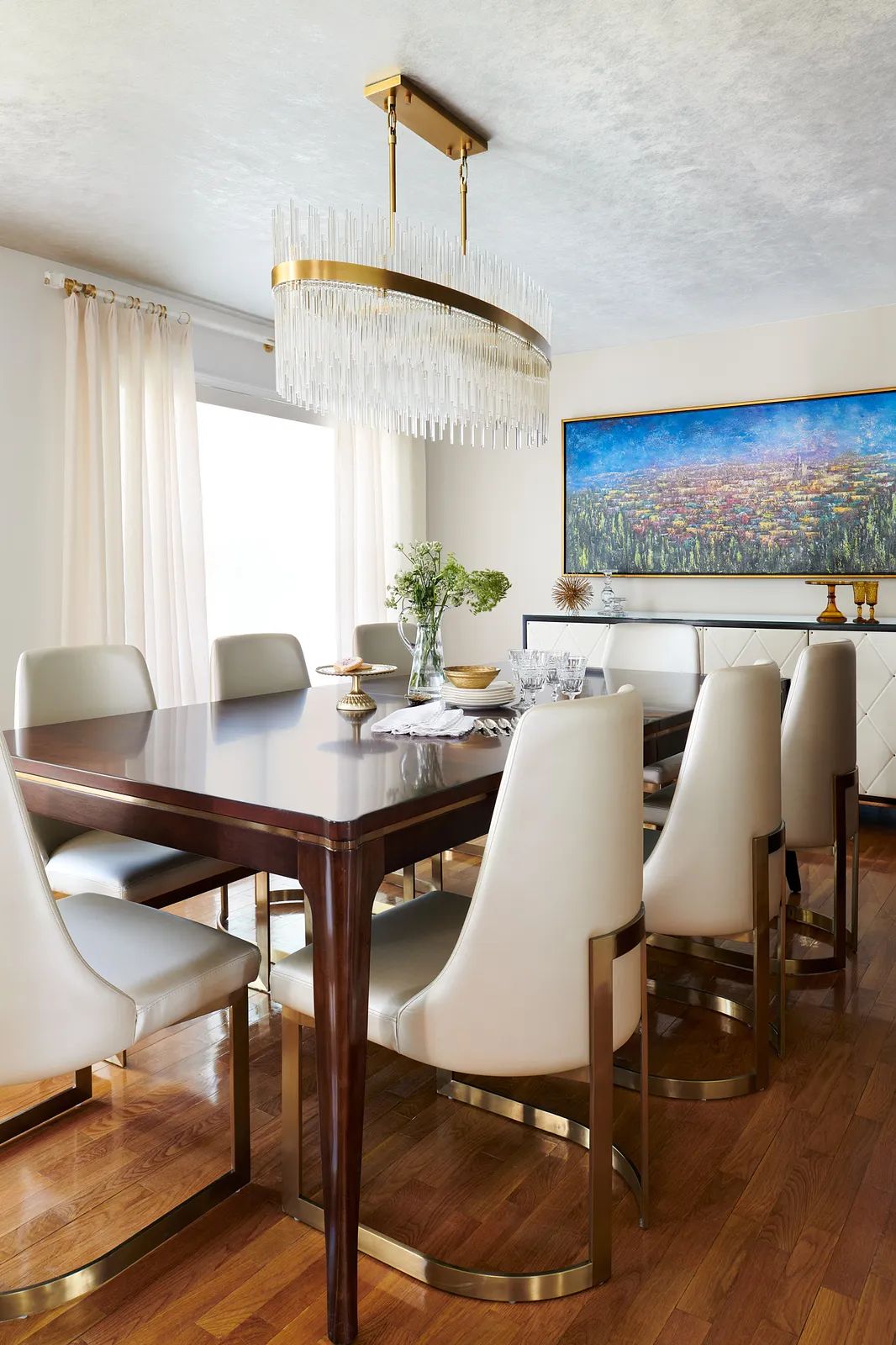 Dining room with wood table, upholstered chairs, large chandelier, curtains, and wall-mounted artwork