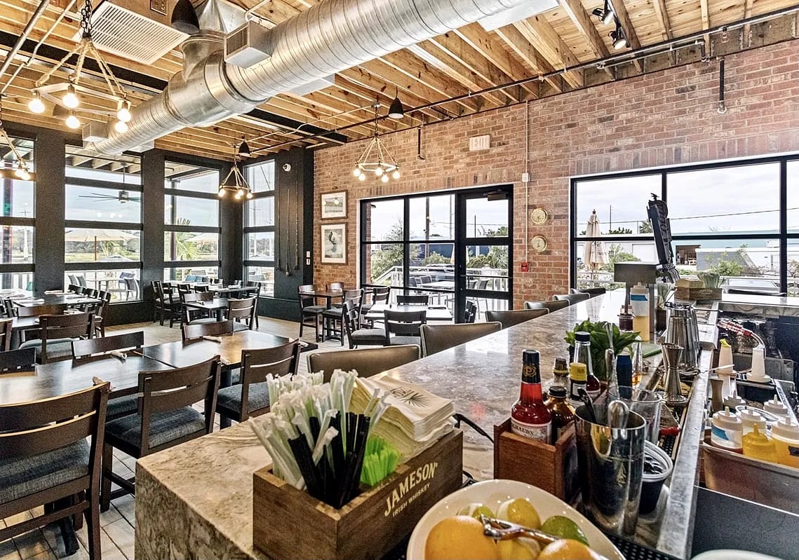Dining room with bar counter, wood table, chairs, large windows, and exposed brick walls.