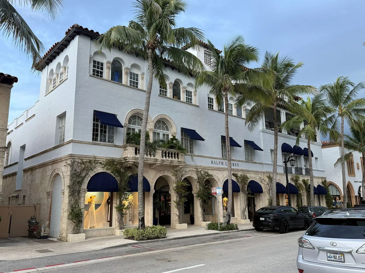 Commercial building with stucco exterior, arched windows, blue awnings, and palm trees.