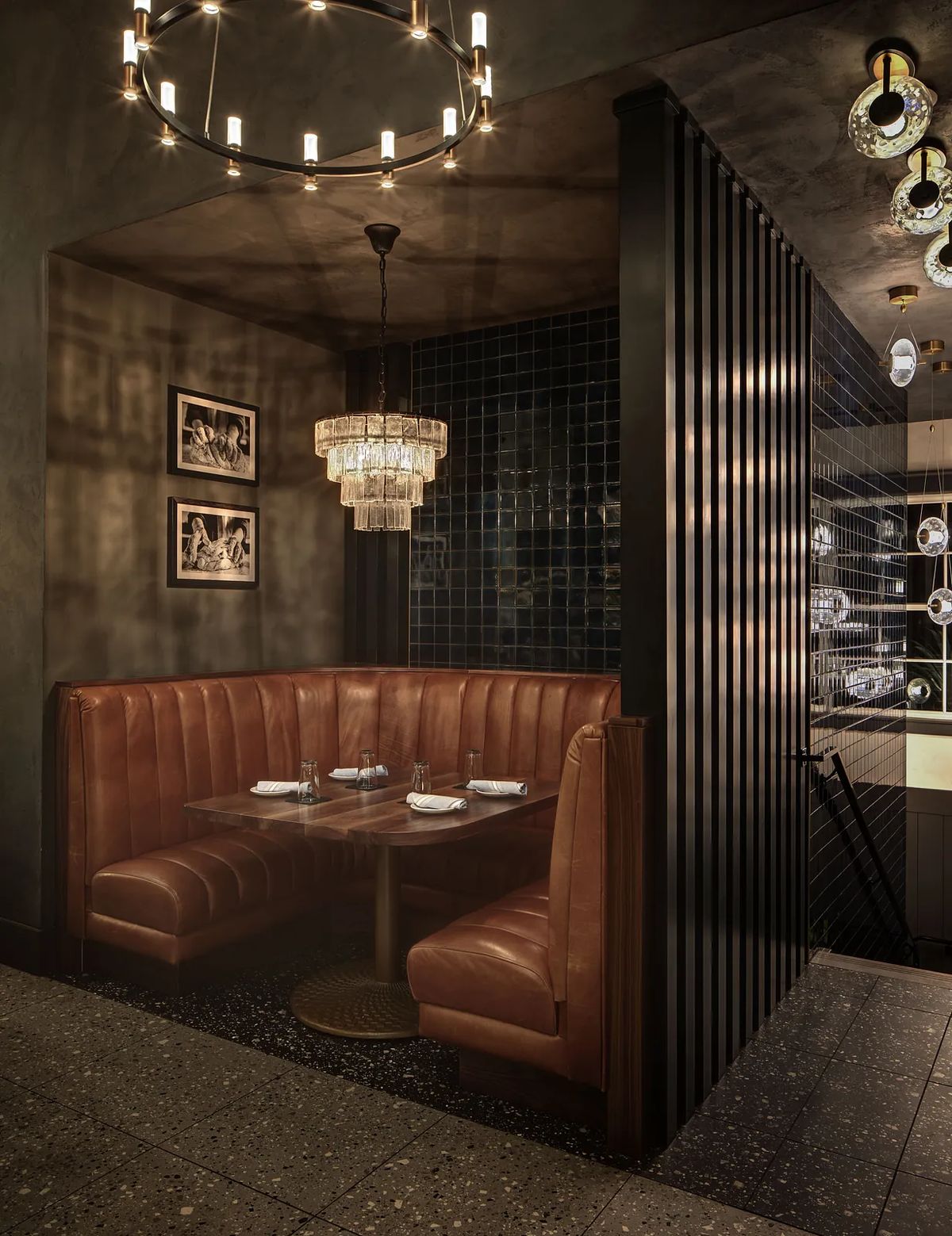Dining room with leather banquette, round table, set diningware, dark tiles, and chandelier.