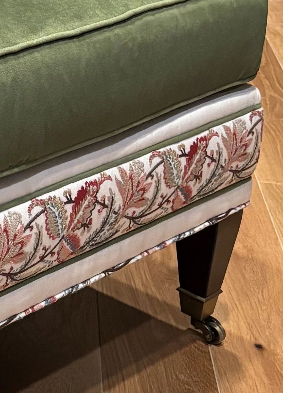Close-up of a green upholstered ottoman with floral embroidery and dark wooden legs.