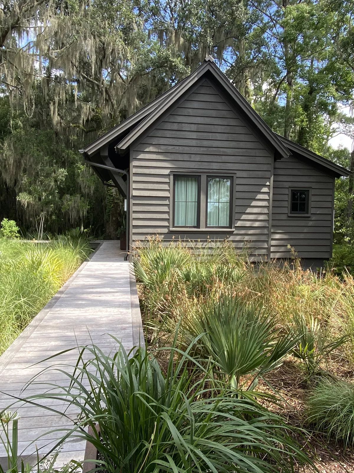 Exterior front of a small wooden house with gray siding, windows, wooden pathway, and surrounding greenery.