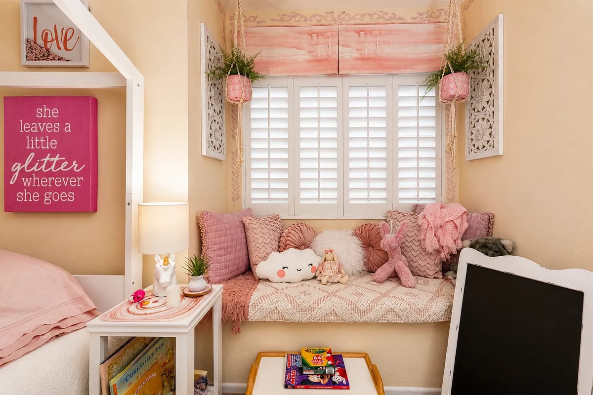 Bedroom corner with bed, nightstand, window seat, cushions, and decorative wall sign.