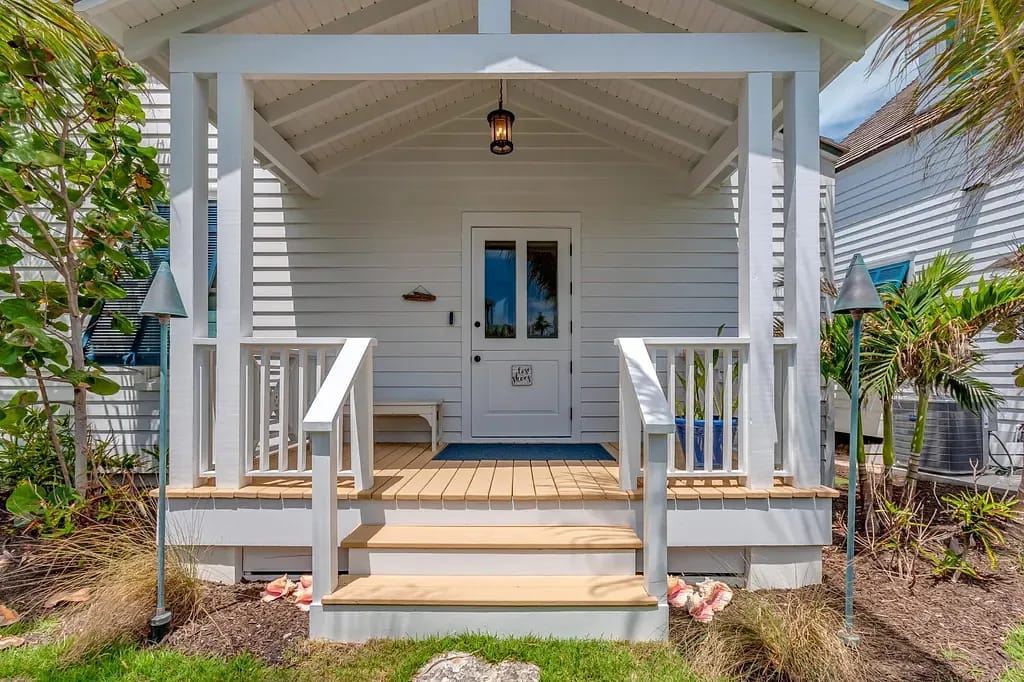 Exterior front with porch, white railing, blue mat, plants, and decorative lamps.