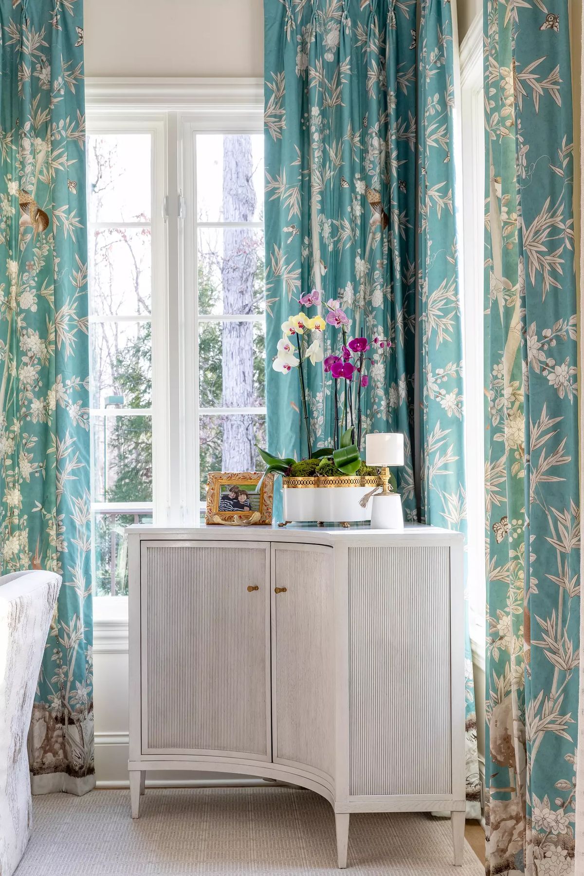 Corner with a light cabinet, floral curtains, potted orchids, and a framed picture.