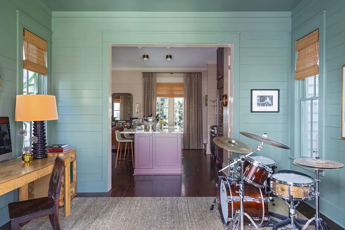 Room with green shiplap walls, wooden desk, drum set, large window, and views into a kitchen area.
