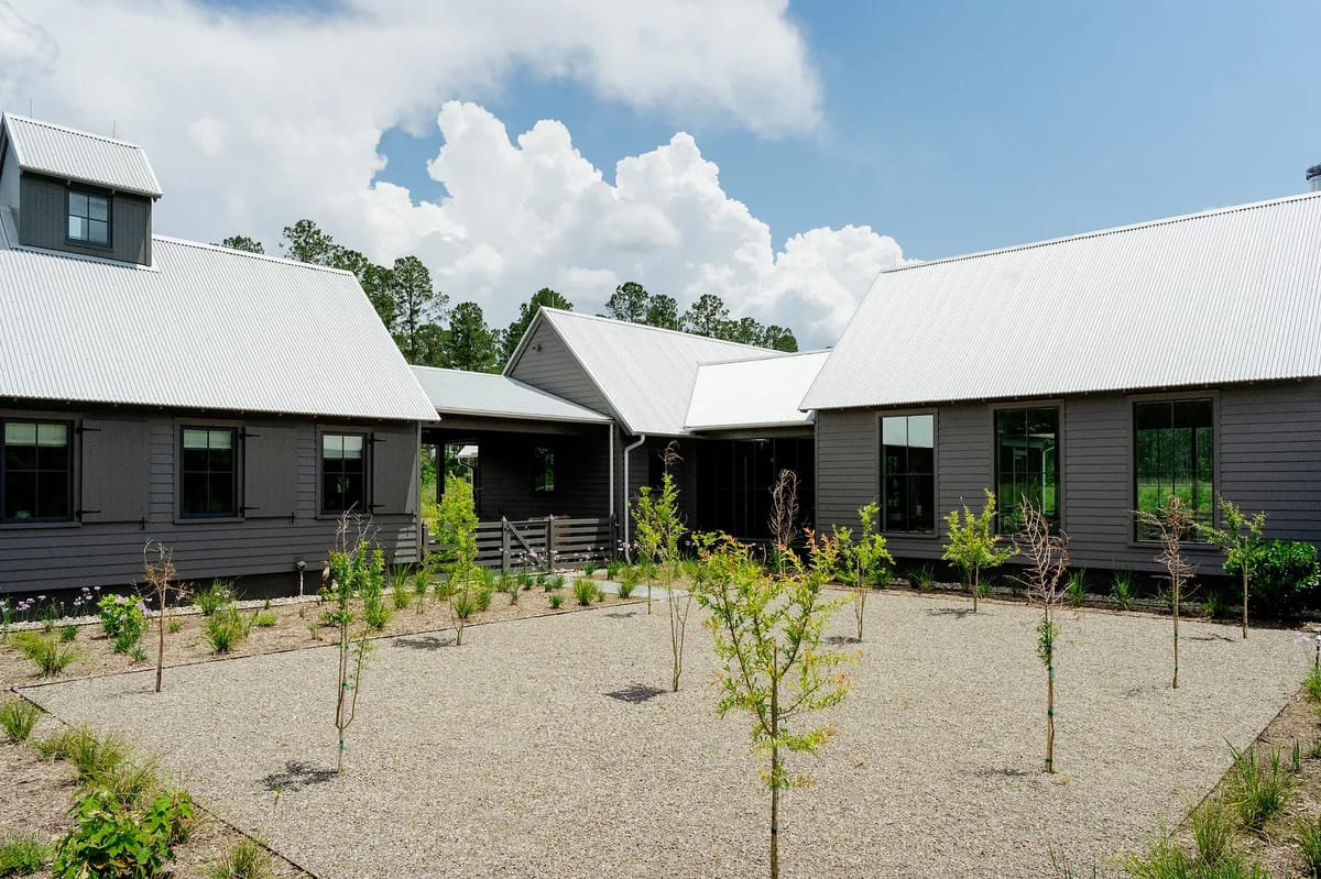 Exterior front view of a house with peaked roofs, gray siding, large windows, and landscaped gravel areas.