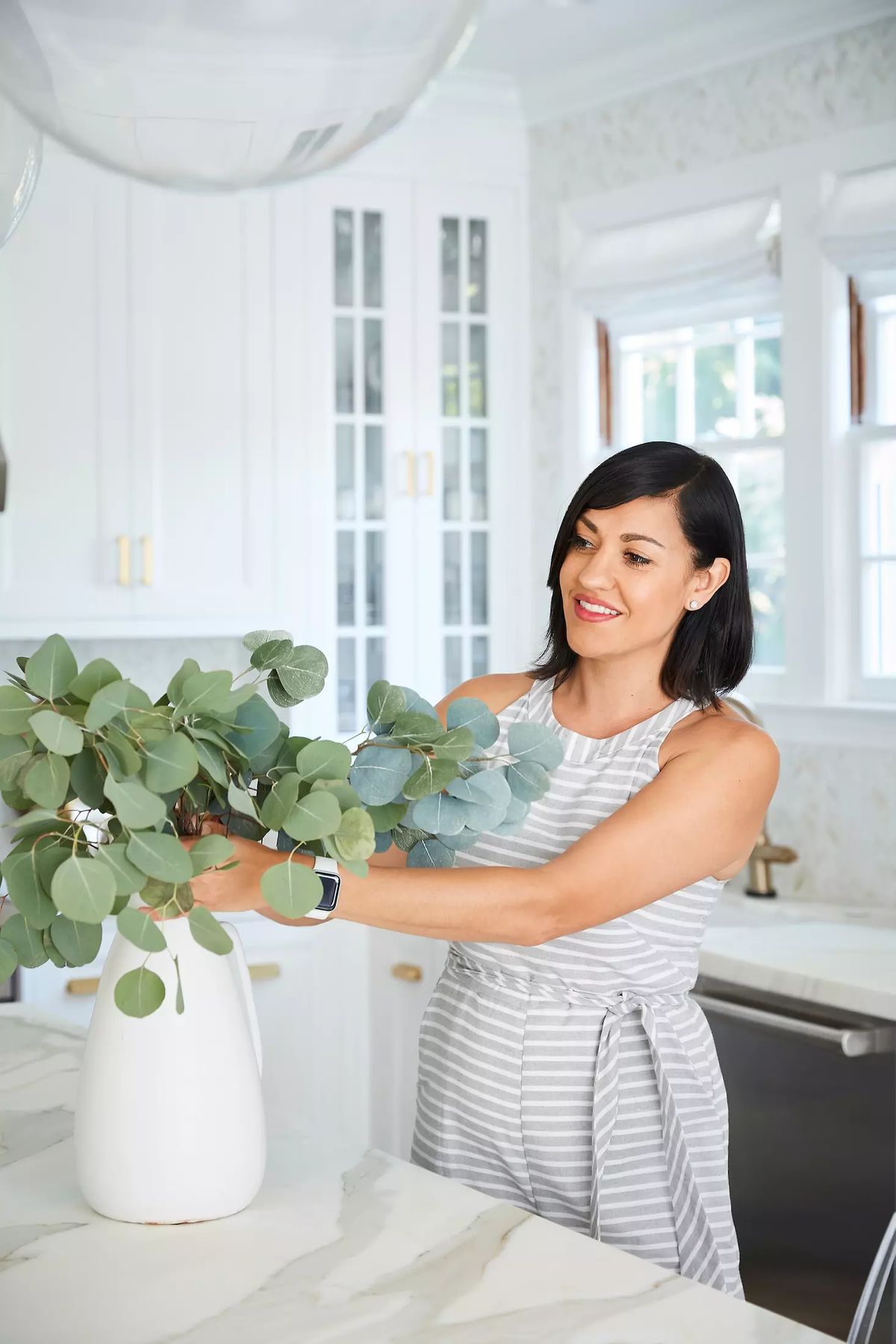Kitchen with marble countertop, white cabinets, large windows, and eucalyptus in a white vase.