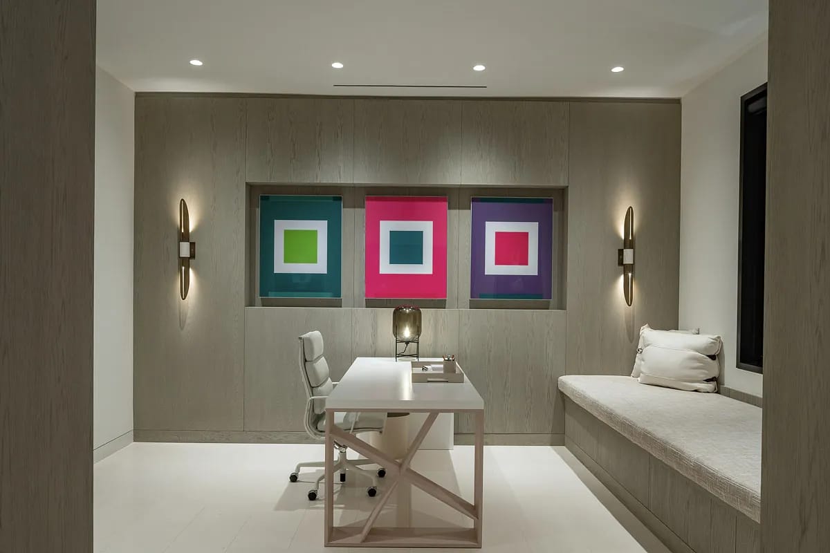 Office with white desk, ergonomic chair, built-in bench, colorful wall art, and light wood paneling