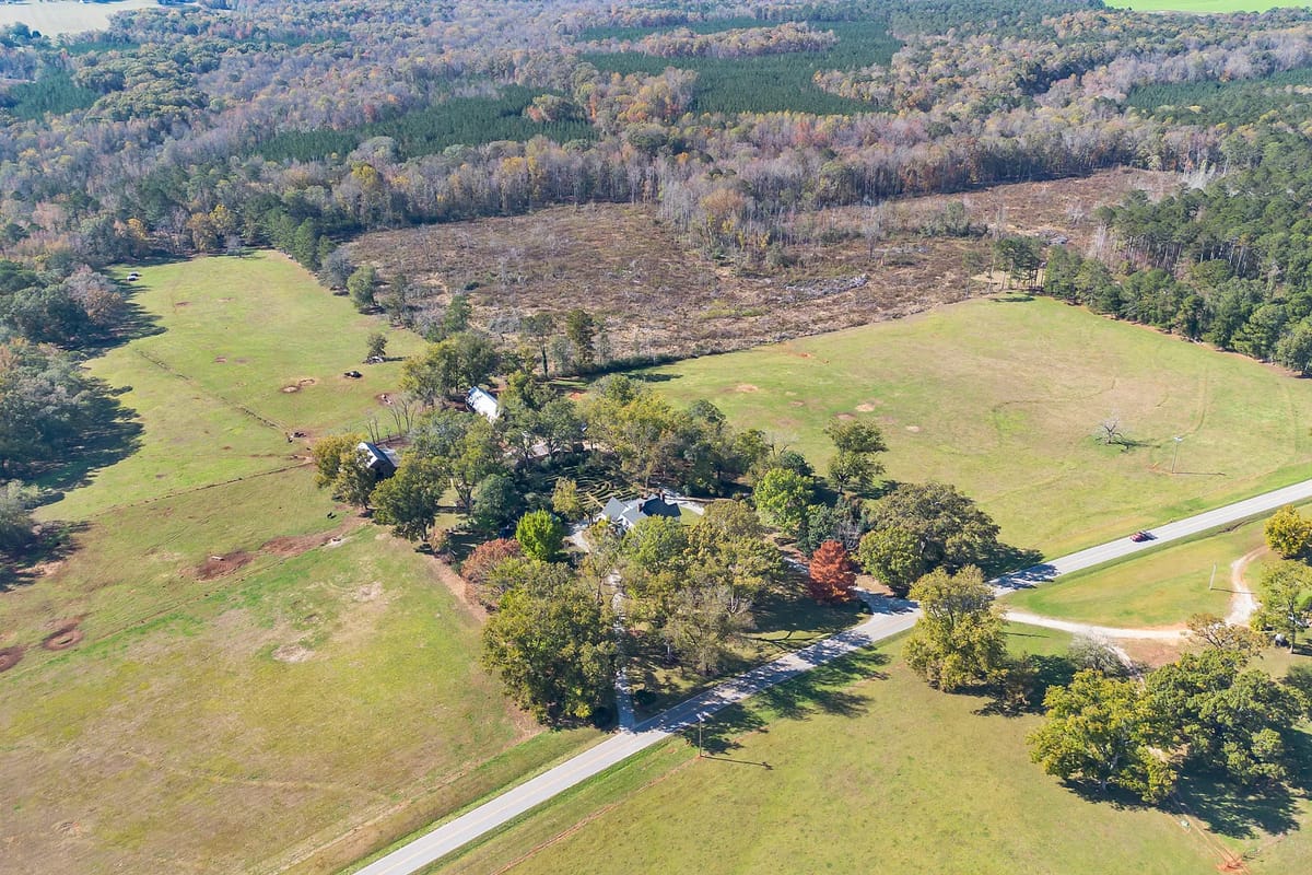 Aerial view of rural landscape with fields, trees, dirt road, and cluster of buildings.