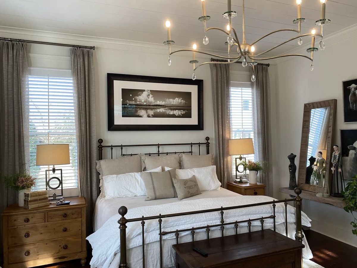 Bedroom with metal bed frame, white bedding, bedside tables, lamps, artwork, and gray curtains.