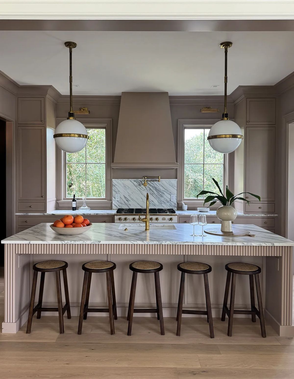 Kitchen with marble island, wooden stools, gray cabinetry, two pendant lights, and large windows.