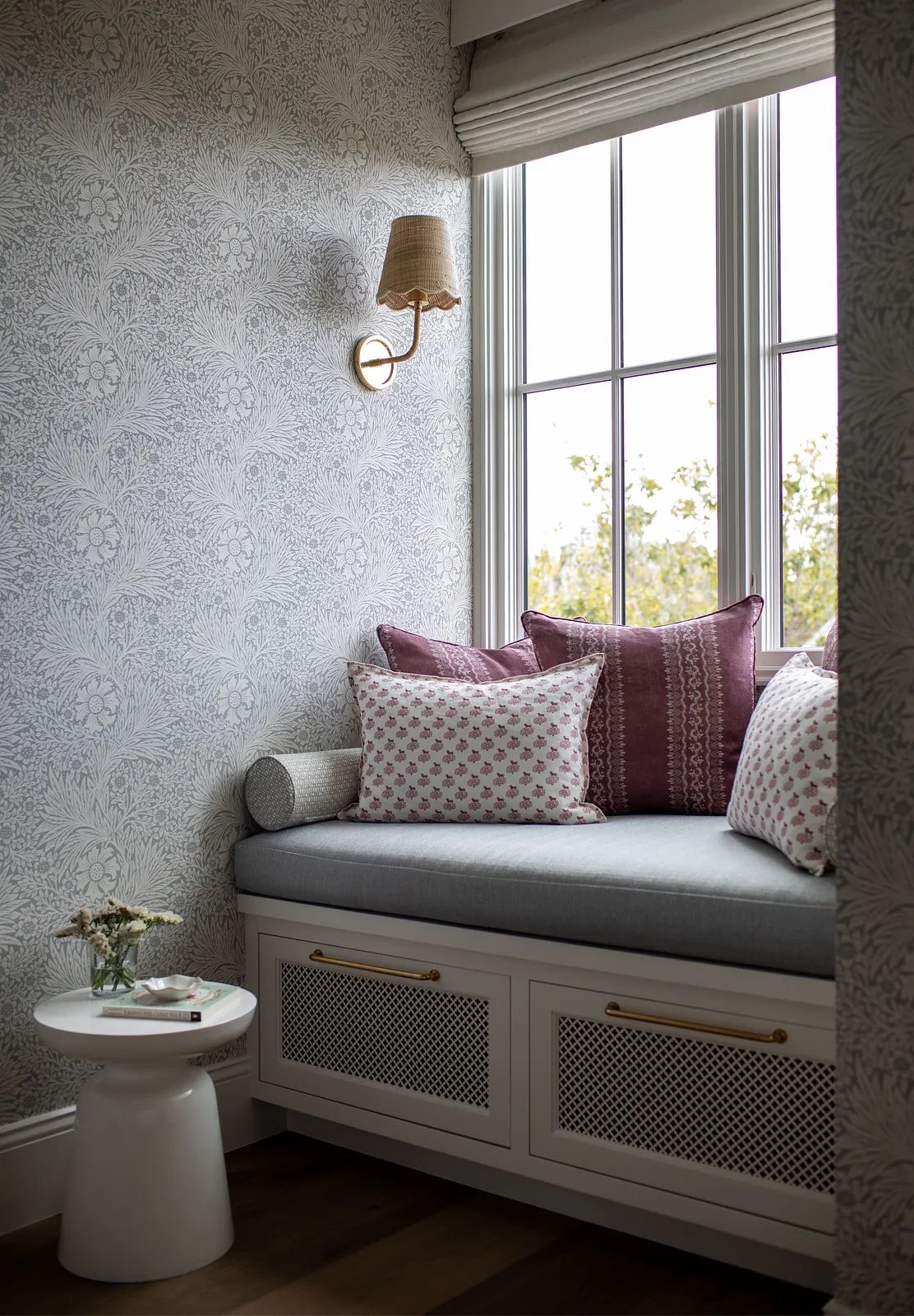 Corner nook with built-in bench, gray cushions, decorative pillows, small table, and floral wallpaper.