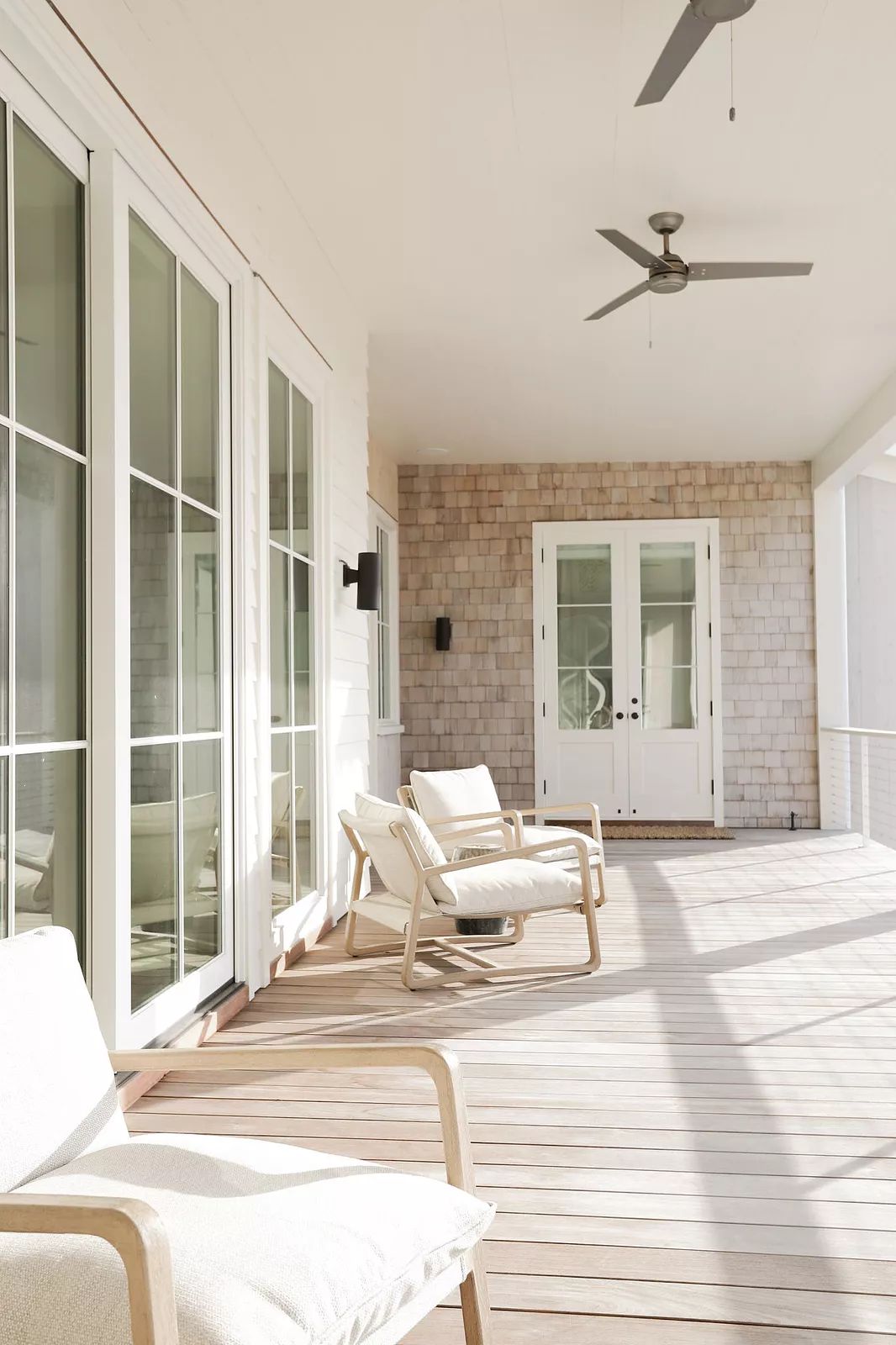 Wooden deck with two armchairs, ceiling fan, and large glass doors leading to a house.