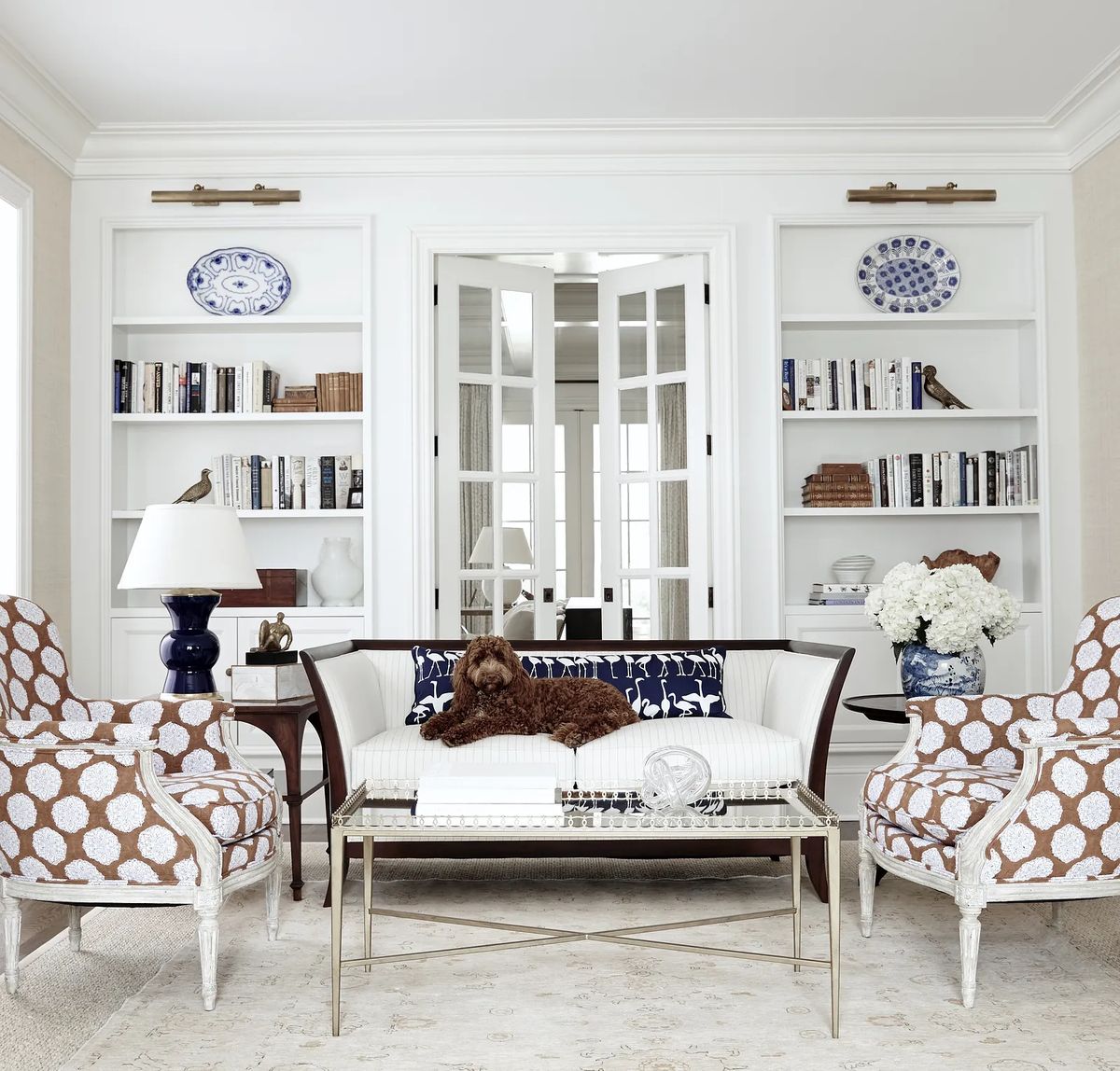 Living room with white settee, patterned chairs, glass coffee table, bookshelves, and a brown dog.